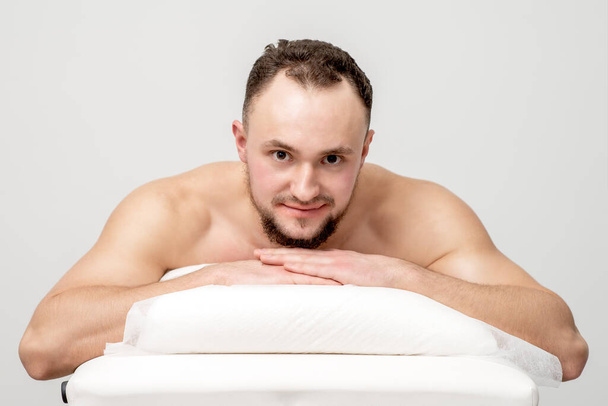 Молодой кавказский мужчина лежит на столе спа, ждет косметических процедур и смотрит в камеру - Фото, изображение