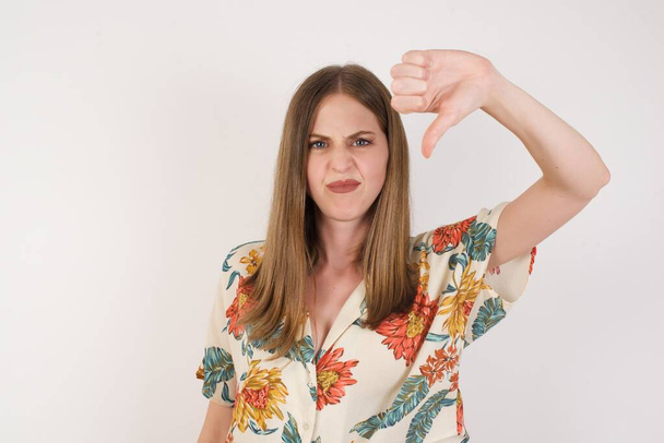 Discontent European woman shows disapproval sign, keeps thumb down, expresses dislike, frowns face in discontent, dressed in white shirt, isolated over gray background. Body language concept. - Photo, Image