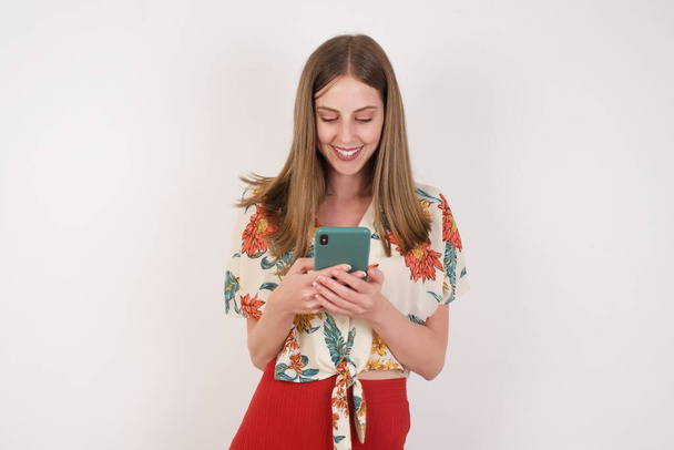 Blond glimlachende jonge vrouw demonstreren witte tanden met behulp van mobiele telefoon, berichten, blij zijn om te sms 'en met haar vriendje, kijken naar het scherm van de smartphone. Moderne technologieën en communicatie - Foto, afbeelding