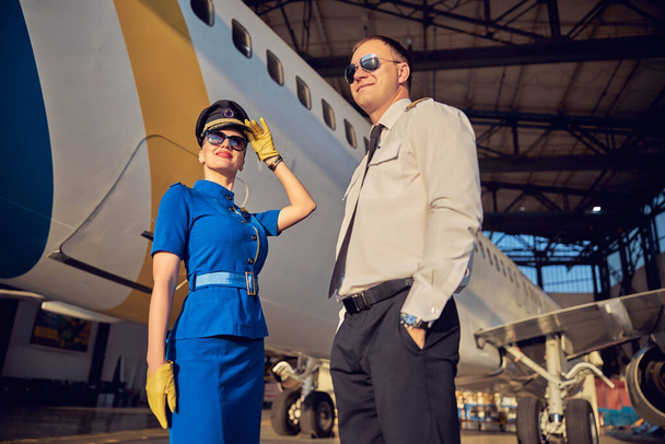 Feliz sorridente mulher e homem vestindo uniforme de negócios em pé na garagem da aviação - Foto, Imagem
