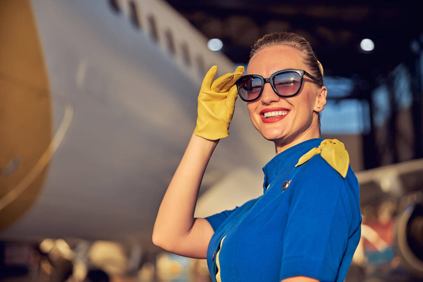 Очаровательная стюардесса в соответствующей форме и кожаных перчатках стоит на открытом воздухе в солнечный день - Фото, изображение