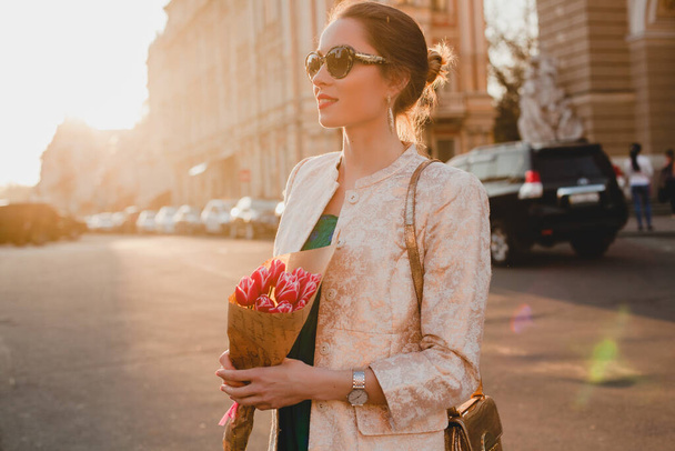 young stylish beautiful woman walking on city street on sunset, europe vacation, holding tulips bouquet, flowers, spring trend, sunglasses, smiling, luxury, red lips, fashionable outfit, jacket - Zdjęcie, obraz
