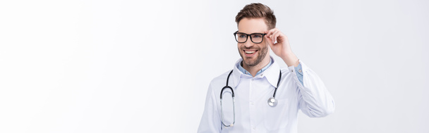 Happy doctor with stethoscope, holding eyeglasses frame, while looking away isolated on white, banner - Foto, Imagem