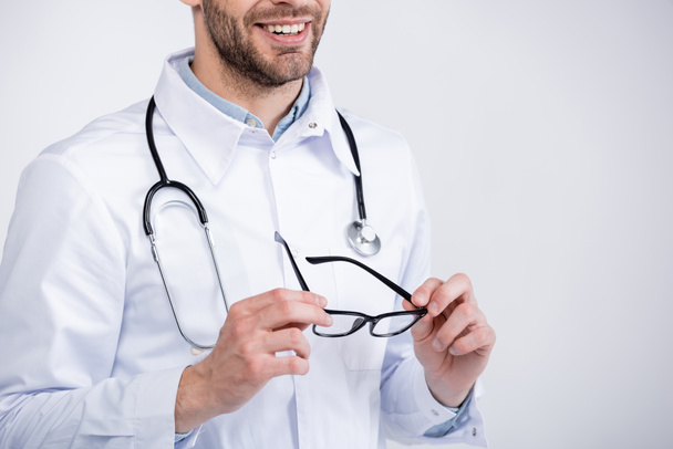 Άποψη χαμογελαστή οφθαλμίατρος με στηθοσκόπιο κρατώντας γυαλιά που απομονώνονται σε λευκό - Φωτογραφία, εικόνα