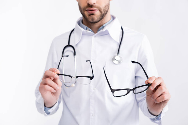 Vista recortada del médico en bata blanca sosteniendo par de anteojos aislados en blanco - Foto, Imagen