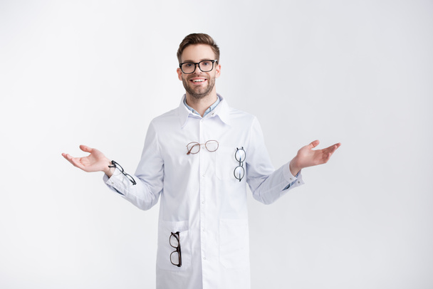 Вид спереди улыбающегося офтальмолога с открытыми руками в белом халате с висящими парами очков, изолированных на белом - Фото, изображение