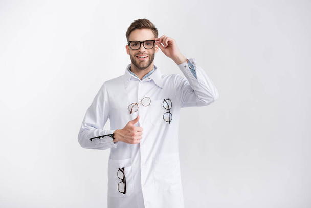 Vista frontal de oftalmólogo sonriente con el pulgar hacia arriba, con abrigo blanco con pares de anteojos colgantes aislados en blanco - Foto, Imagen