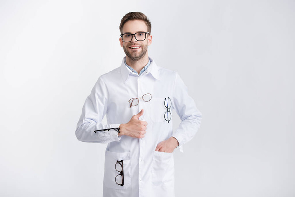 Vista frontal del médico sonriente con el pulgar hacia arriba y la mano en el bolsillo, con abrigo blanco con pares de anteojos colgantes aislados en blanco - Foto, Imagen