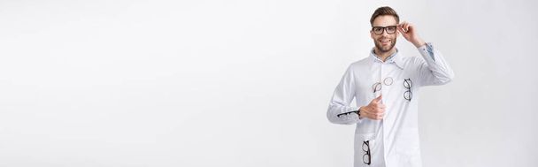 Vista frontal del oftalmólogo feliz con el pulgar hacia arriba, con abrigo blanco con pares de anteojos colgantes aislados en blanco, pancarta - Foto, Imagen