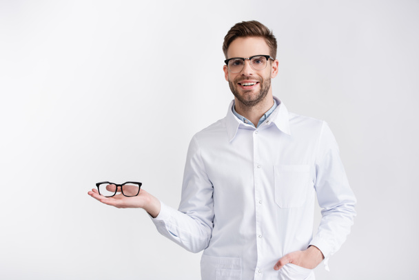 Vooraanzicht van een lachende arts met hand in hand bril op de palm, terwijl hij kijkt naar camera geïsoleerd op wit - Foto, afbeelding