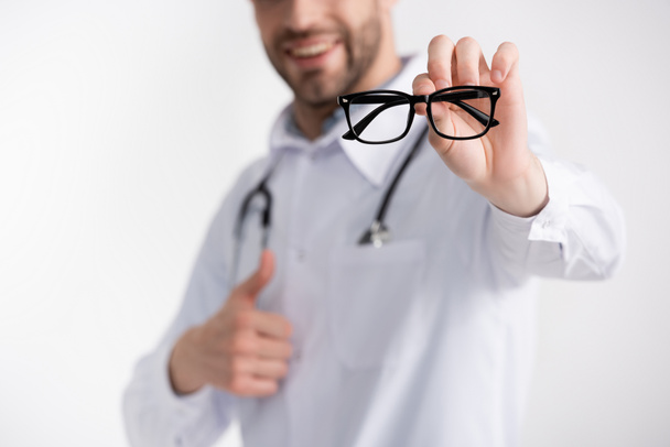 Cropped view of doctor with thumb up, showing eyeglasses with black frame isolated on white on blurred background - Photo, image
