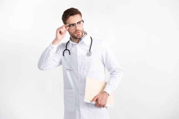 Médico adulto joven con una carpeta que sostiene el marco de las gafas, mientras mira hacia otro lado aislado en blanco - Foto, Imagen