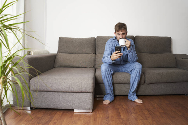 Νεαρός άνδρας ντυμένος με πιτζάμες στο σπίτι πίνοντας καφέ κάθεται στον καναπέ ενώ ελέγχει το smartphone. - Φωτογραφία, εικόνα
