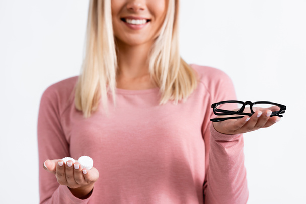 Gehakt zicht op bril en contactlenzen in handen van lachende vrouw op wazige achtergrond geïsoleerd op wit - Foto, afbeelding