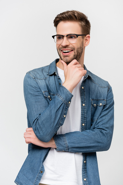 Mladý muž v brýlích se usmívá a dotýká se brady izolované na šedé - Fotografie, Obrázek
