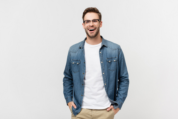 Positive man in eyeglasses holding hands in pockets of pants and looking away isolated on grey - Foto, afbeelding