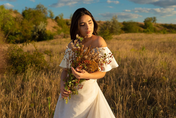 young beautiful girl in a white dress with wildflowers, in the field. Against the backdrop of a cloudy blue sky. Wedding dress, the bride stands with flowers in the field. Wedding dress of the bride. Young girl twenty two years old in a white dress,  - Foto, Bild