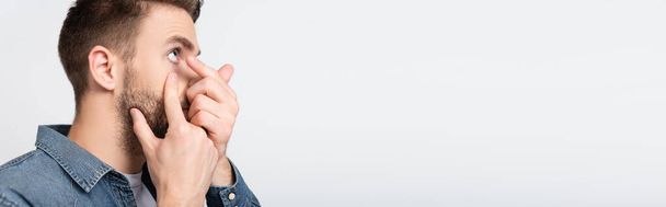 Mężczyzna dotykający policzka nosząc soczewkę kontaktową odizolowaną na szarym, banerowym  - Zdjęcie, obraz