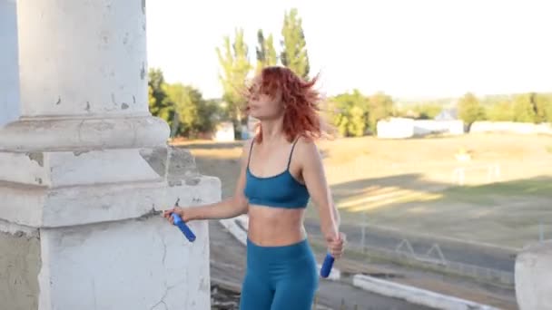 Genç kızıl saçlı atlet kız sokakta ip atlıyor. Aktif yaşam tarzı. Tozluklu atletik bir kadın vücudu ve turkuaz bir bluz, beyaz spor ayakkabılar, havaya zıplayan. - Video, Çekim