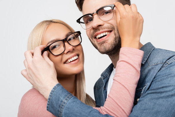 Usmívající se pár dívá na fotoaparát při dotyku brýle na sebe izolované na šedé - Fotografie, Obrázek