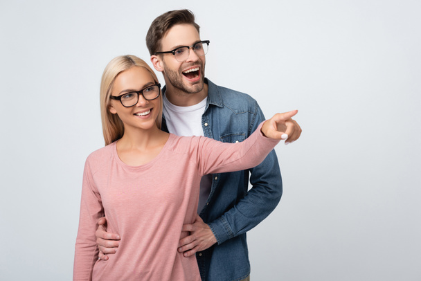 Cheerful man in eyeglasses embracing girlfriend pointing with finger isolated on grey - Foto, Imagem