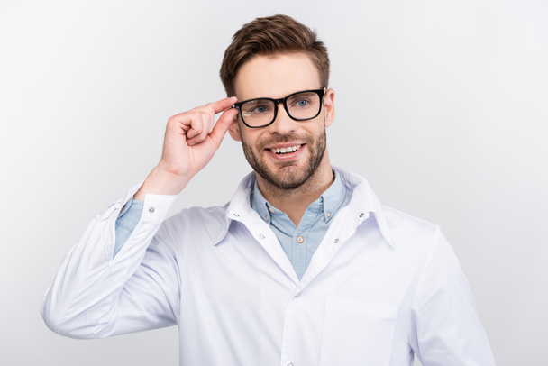 Lächelnder Arzt im weißen Kittel berührt Brille isoliert auf grau  - Foto, Bild