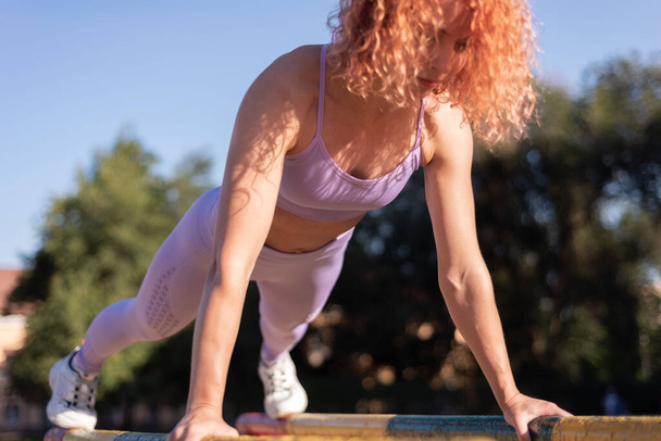 young red-haired sportswoman doing push-ups on the uneven bars on the street. In pink sportswear. active lifestyle, sports style Fitness woman doing push-ups on uneven bars in crossfit gym. Workout lifestyle concept - Photo, Image