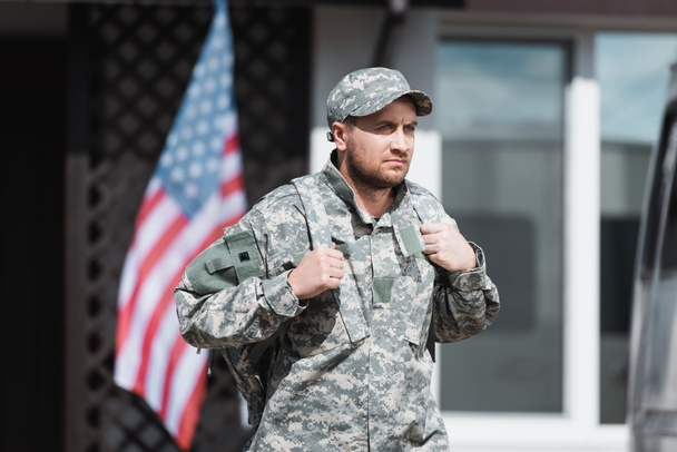 Серйозний військовий чоловік у камуфляжі дивиться в сторону з розмитим будинком і прапором США на фоні
 - Фото, зображення