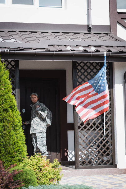 Grave uomo militare in piedi vicino alla porta della casa e guardando lontano vicino cespugli e bandiera americana - Foto, immagini