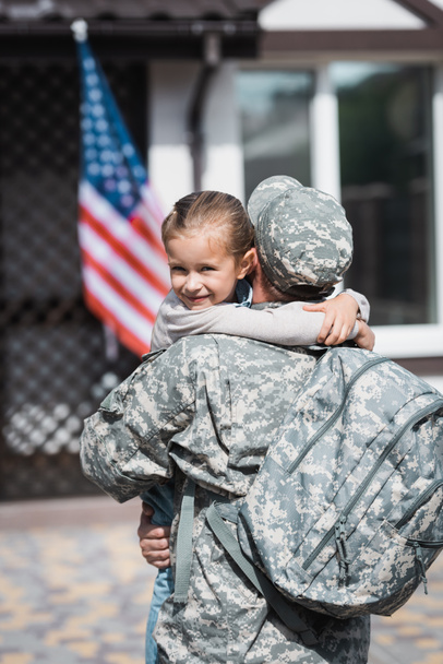 Vista trasera del militar con la mochila abrazando a la hija con la bandera americana borrosa y la casa en el fondo - Foto, Imagen