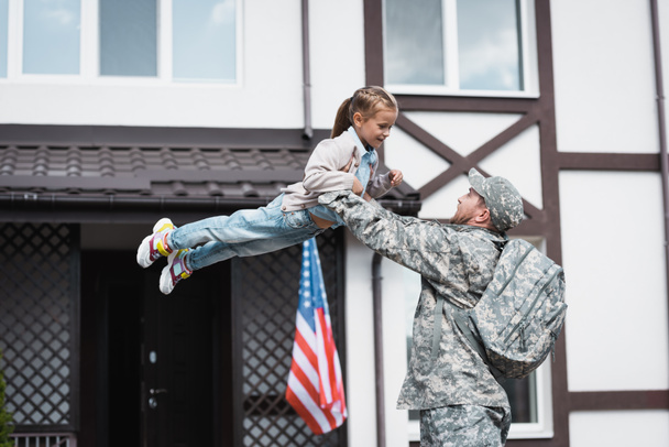 Military man lifting smiling daughter in air near house with american flag - Photo, Image