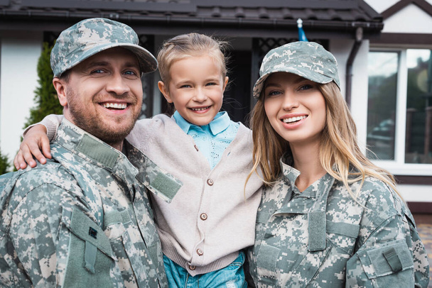 Šťastný otec a matka ve vojenských uniformách zvedá dceru a dívá se na fotoaparát na rozmazaném pozadí - Fotografie, Obrázek