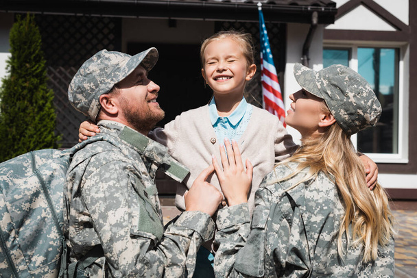Szczęśliwa córka obejmująca ojca i matkę w mundurach wojskowych w pobliżu domu z flagą USA - Zdjęcie, obraz
