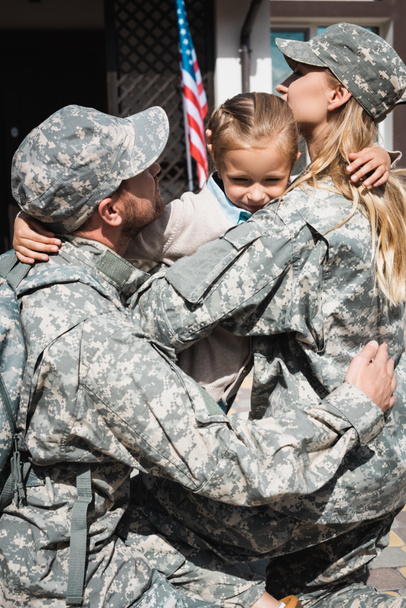 Zdenerwowana córka przytulająca matkę i ojca w wojskowych mundurach w pobliżu domu z amerykańską flagą - Zdjęcie, obraz