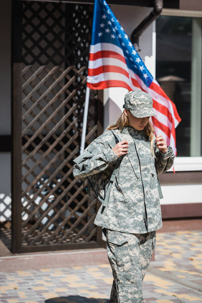Soldatin verlässt Haus und hält Rucksack-Schultergurte mit US-Flagge im Hintergrund - Foto, Bild