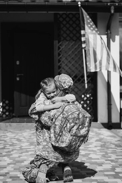 Military servicewoman embracing daughter, while sitting on knee near house and american flag, monochrome - Photo, Image