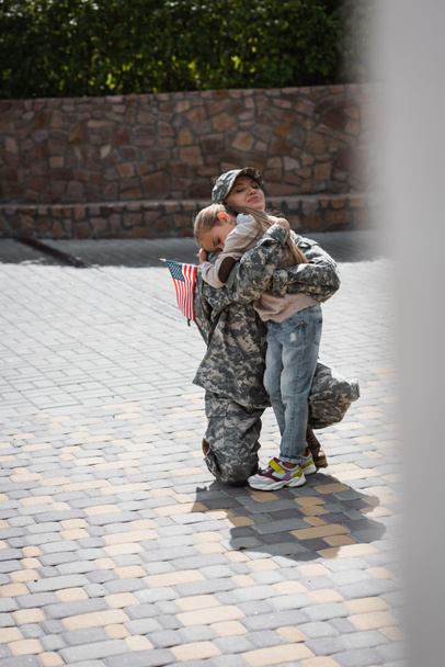 Lachende moeder in militair uniform zittend op knie en omhelzende dochter op wazige voorgrond - Foto, afbeelding