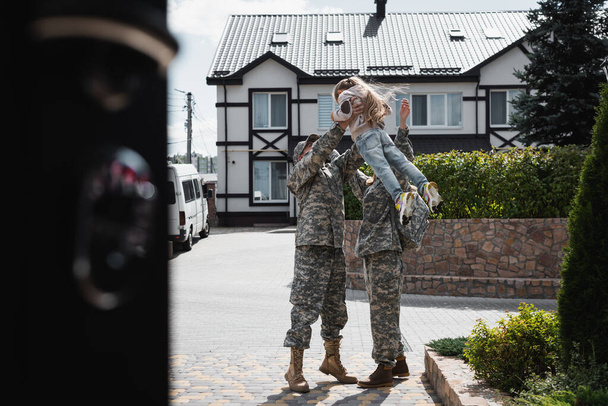 軍の制服を着た父と母が娘を空中に持ち上げながら、前景がぼやけて通りに立っている - 写真・画像