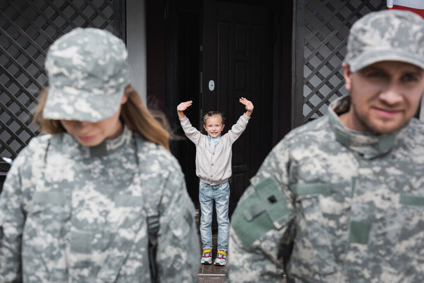 Menina sorridente com as mãos acenando em pé perto da porta da casa com homem e mulher desfocados em uniformes militares em primeiro plano - Foto, Imagem