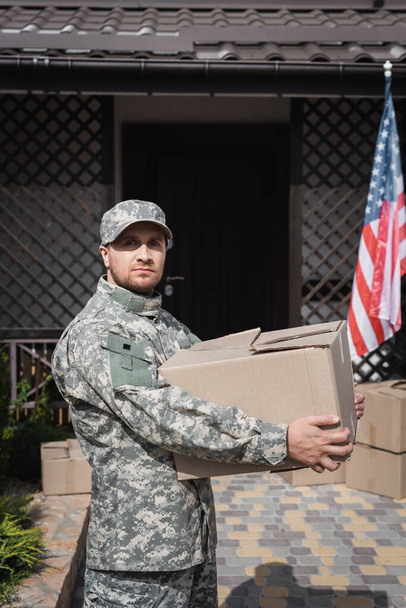 Військовослужбовець тримає картонну коробку, дивлячись на камеру біля будинку та прапор Америки. - Фото, зображення