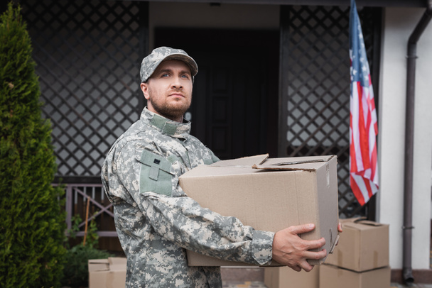 Military serviceman holding cardboard box, while looking away near house and american flag - Photo, Image