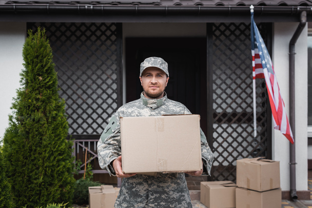 Vue de face du militaire avec boîte en carton debout près de la maison avec drapeau américain - Photo, image
