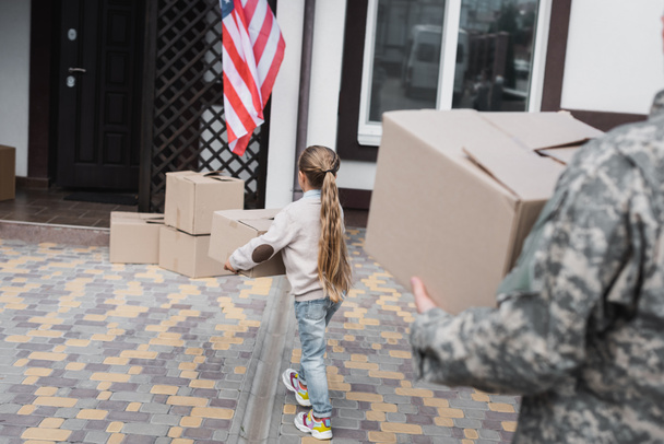 Back view of girl carrying cardboard box near house with blurred father on foreground - Photo, Image