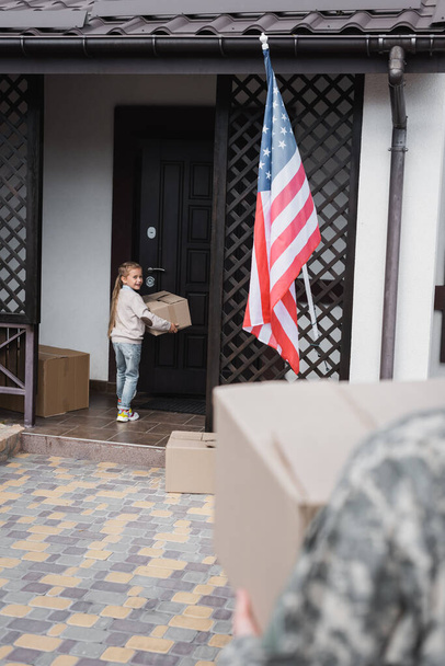 Menina sorridente com caixa de papelão em pé perto da porta da casa com pai desfocado em primeiro plano - Foto, Imagem