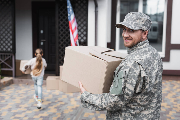 Happy military serviceman holding cardboard box and looking at camera with blurred girl on background - Photo, Image