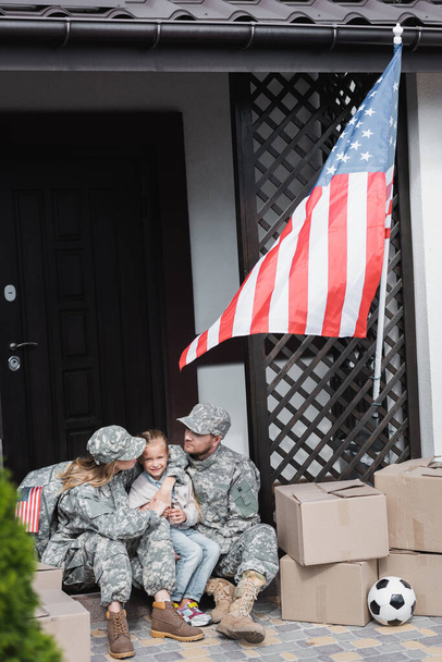 Wojskowi rodzice z przytulaniem córki, siedząc na progu obok kartonowych pudełek i amerykańskiej flagi na niewyraźnym pierwszym planie - Zdjęcie, obraz