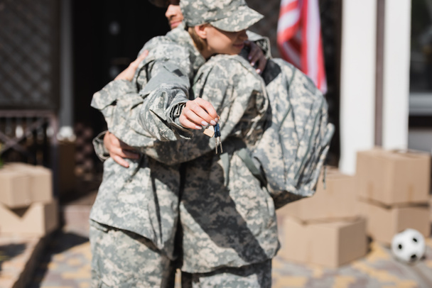 Στρατιωτικό ζευγάρι αγκαλιάζει και κρατώντας τα κλειδιά με θολό σπίτι και κουτιά από χαρτόνι στο παρασκήνιο - Φωτογραφία, εικόνα