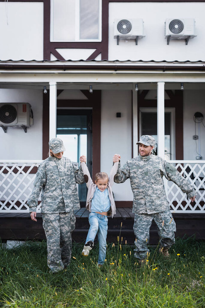 Güler yüzlü asker baba ve anne kızı evin yakınındaki çimlerin üzerinden kaldırıyor. - Fotoğraf, Görsel