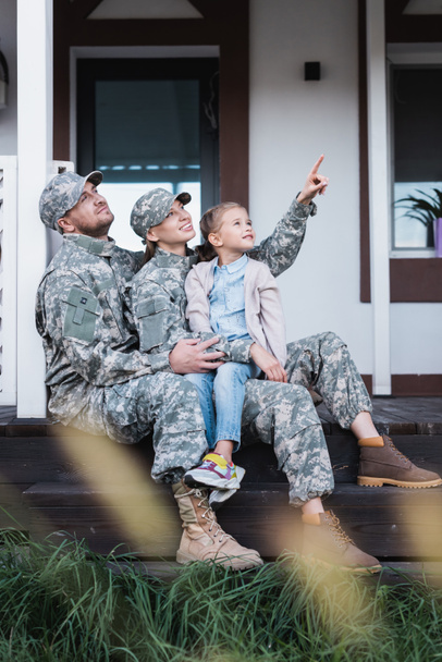 Heureux père et mère militaires pointant du doigt, regardant ailleurs, assis avec sa fille sur le seuil de la maison au premier plan flou - Photo, image