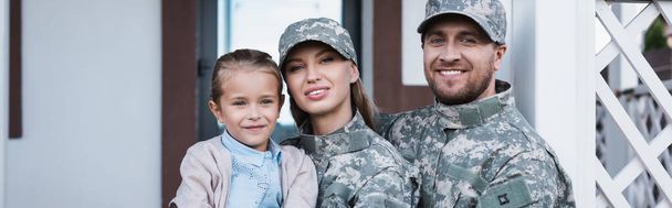 Porträt einer lächelnden Militärmutter und eines Vaters mit Tochter, die in die Kamera mit Haus im Hintergrund blicken, Banner - Foto, Bild